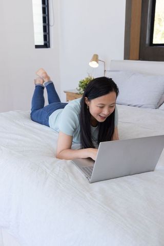 Asian woman reclining on bed using laptop for remote work and study in minimalist bedroom