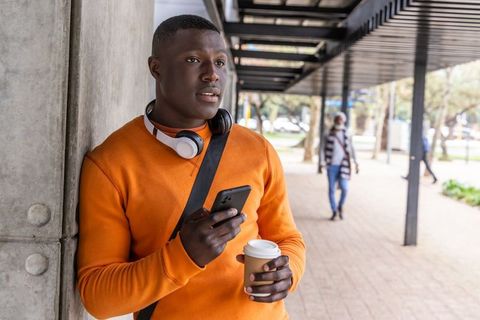 African american man leaning on wall using smartphone with headphones holding coffee cup