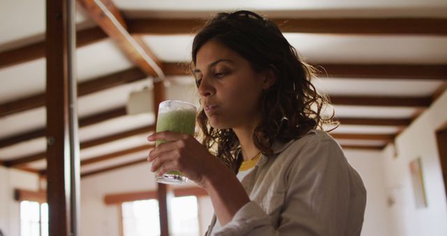 Woman enjoying a freshly made green smoothie in a comfortable home setting. Ideal for use in promotions related to health, nutrition, and wellness. Great for articles and campaigns focusing on healthy living, personal well-being, and homemade nutritious drinks.