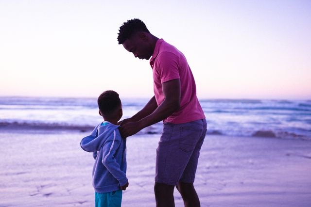 African american young father wearing jacket to son at beach against clear sky at sunset, copy space. nature, unaltered, family, love, togetherness, childhood, enjoyment and holiday concept.