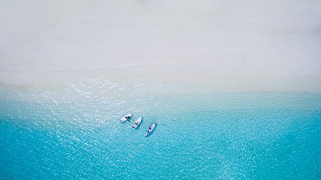 This high-angle view captures a serene scene of people engaging in paddleboarding on crystal clear turquoise waters close to a pristine sandy beach. The image could be used for travel and tourism promotions, outdoor adventure campaigns, tropical destination advertising, or to evoke a sense of tranquility and escape in lifestyle blogs or magazines.