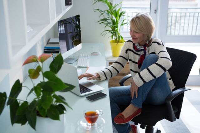 Woman sitting at desk using laptop in home office. Casual and relaxed atmosphere with plants and tea on the table. Ideal for illustrating remote work, freelance lifestyle, productivity, and modern home office setups.
