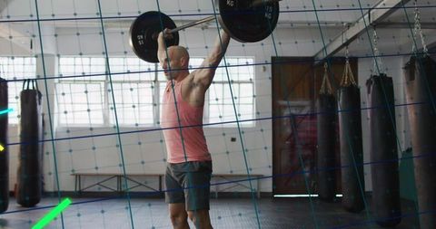 Bald athlete lifting heavy barbell overhead in boxing gym with punching bags and daylight