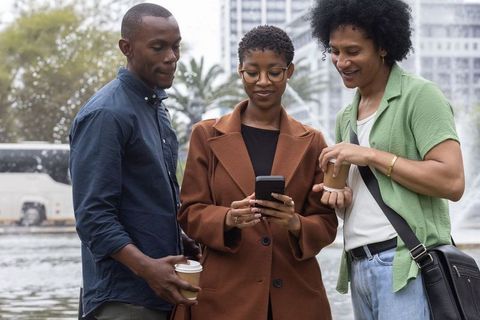 African american friends checking smartphone while holding to-go coffee by city fountain