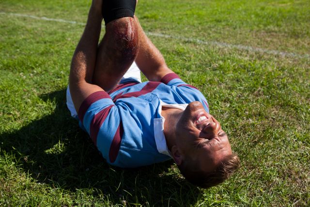 High angle view of rugby player with injured knee lying on field during sunny day