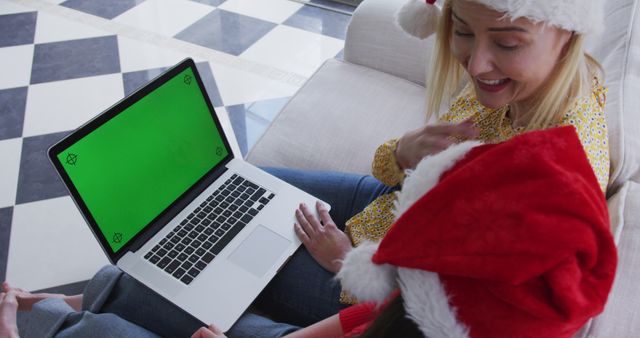 Mother and child seated on sofa using a laptop with green screen. Both wearing Christmas Santa hats signifies holiday season celebration. Perfect for depicting family bonding, festive tech usage, holiday digital interactions, seasonal ads, or family-oriented technology services during Christmas.