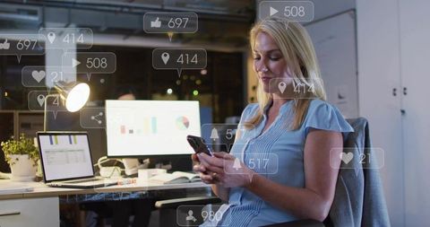 Woman scrolling smartphone in modern office at night with analytics screens and overlays