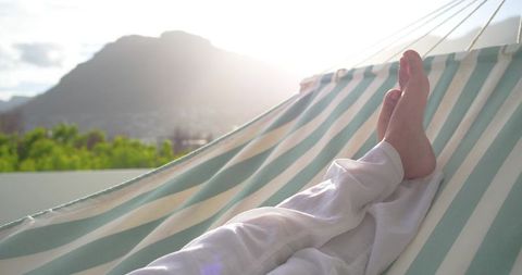 Indian adult male reclining in sunlit striped hammock on balcony with mountain view