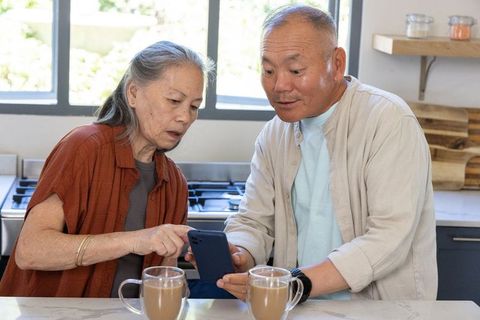 Asian senior couple sharing smartphone at kitchen counter with coffee mugs smiling