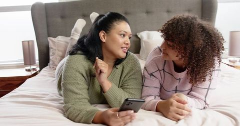 Female Friends Lying on Bed Wearing Cozy Sweaters Holding Smartphone Chatting in Sunlit Bedroom