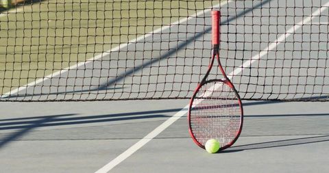 Red tennis racket leaning against net with yellow ball on court lines and long shadows