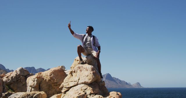 Man standing confidently on rocky cliff near the sea taking selfie. Ideal for use in travel promotions, adventure posters, tourism campaigns, and outdoor activity blogs. Great depiction of exploring nature and capturing memorable moments.