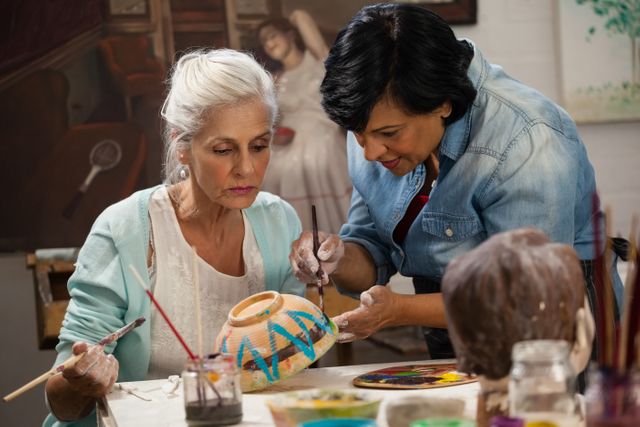 This image captures a moment of mentorship and creativity in an art class, where a woman is assisting a senior woman in painting a bowl. It is ideal for use in content related to art education, senior activities, creative workshops, and intergenerational learning. The image highlights the importance of hands-on activities and the joy of artistic expression.