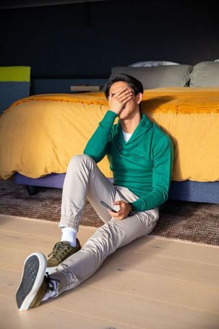 Man sitting on floor leaning against bed covering face holding phone stress fatigue