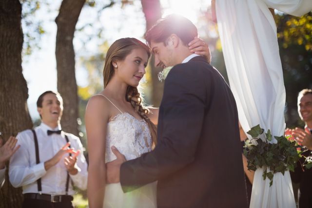 This image shows a romantic moment between a bride and groom embracing during their outdoor wedding ceremony. The sunlight filters through the trees, adding a magical touch to the scene. Guests are seen clapping in the background, celebrating the couple's special day. This image is perfect for use in wedding invitations, romantic greeting cards, wedding planning websites, or any content related to love and marriage.