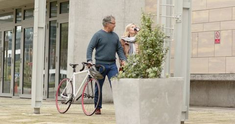 Middle-aged couple walking hand in hand with bicycle and takeaway coffee in urban plaza
