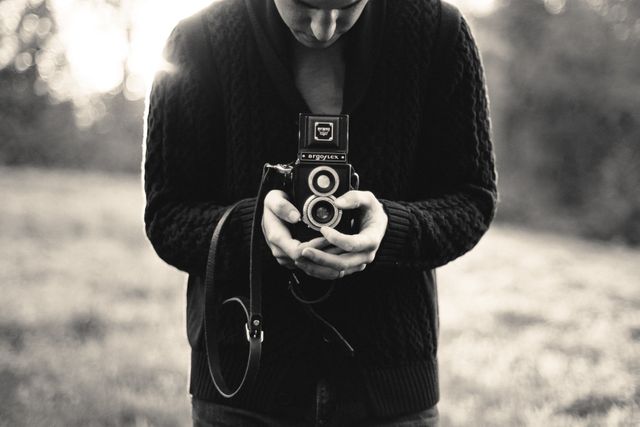 Person using a vintage camera outdoors under natural sunlight. Black and white tones emphasize analog and nostalgic feel. Could be used for concepts such as photography enthusiasts, outdoor adventures, analog film revival, and vintage clothing.