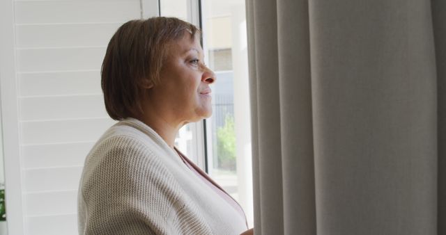 A mature woman is standing near a window, looking outside thoughtfully. She appears to be deep in contemplation while dressed in casual indoor clothing. This image could be used in articles or websites focusing on personal reflection, self-care, peaceful moments at home, or lifestyle themes for older adults.