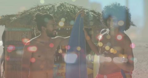 Holding blue surfboard, two men laughing on sunlit sandy beach by thatched hut