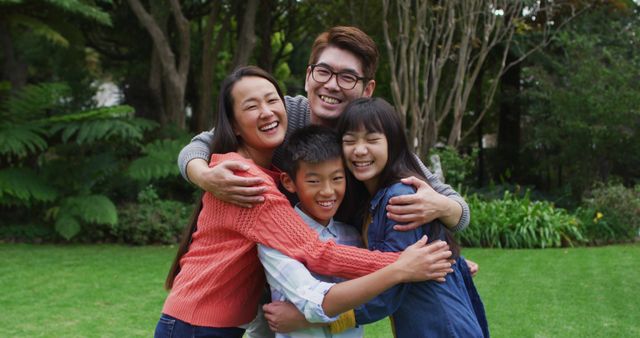 This stock photo features a joyful family of four embracing each other outside in a park setting. Perfect for use in advertisements, social media posts, and blog entries celebrating family values, happiness, and bonding in natural surroundings.