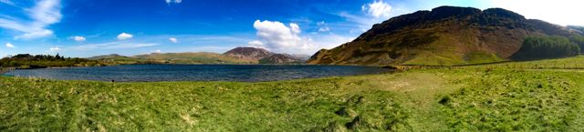 Panoramic view of a tranquil mountain lake surrounded by grassy hills under a bright blue sky with scattered clouds. Perfect for use in travel brochures, nature websites, environmental campaigns, or backgrounds for outdoor adventure promotions.