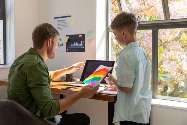 Caucasian dad sitting on his desk in front of his computer with his sob beside him. his son is showing him a drawing.
