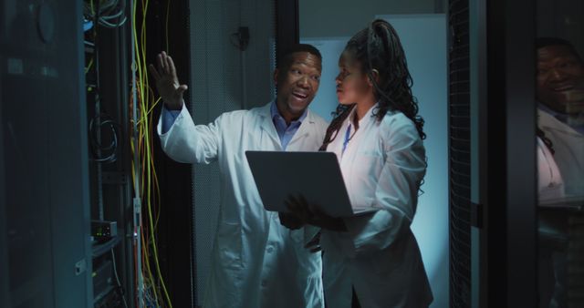 Two IT professionals wearing lab coats having a discussion in a data center filled with equipment and cables. Uses include business, technology, IT industry, engineering teamwork presentations, corporate websites, and educational materials depicting professional work environments.