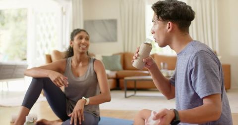 Young man drinking post-workout shake while partner resting and smiling on yoga mat