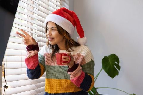 Woman wearing santa hat holding red mug peeking through horizontal blinds in cozy home