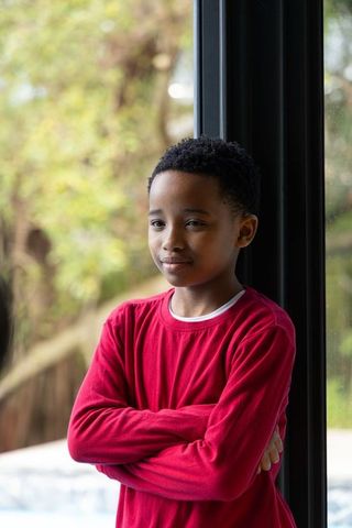 African american preteen standing with arms crossed by window wearing red long-sleeve shirt