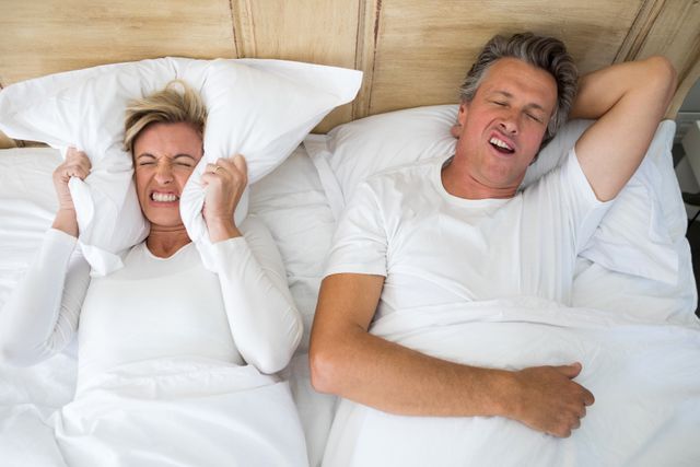 Annoyed woman covering ears with pillow while man snoring on bed in bedroom