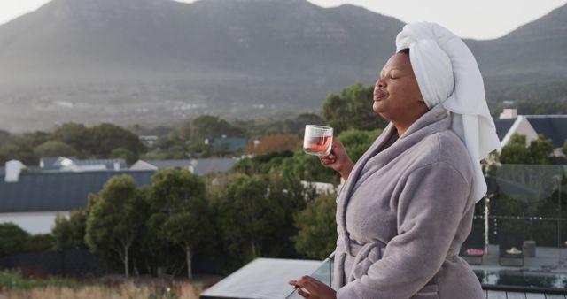 Middle-aged African American woman enjoying morning tea in bathrobe with serene mountain scenery. Great for concepts of relaxation, self-care, peaceful lifestyle, wellness retreats, and morning routines.