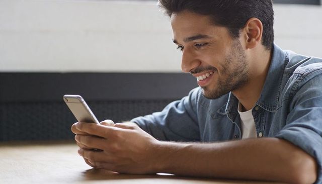 Young man reclining on wooden floor texting and smiling in denim shirt and white tee