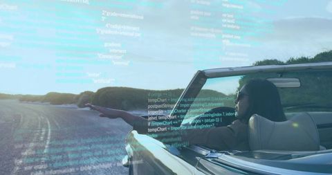 Indian woman driving convertible on coastal road at sunset with digital code overlay