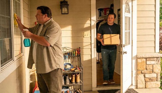 Man cleaning window while companion carrying moving box into sunlit suburban front porch