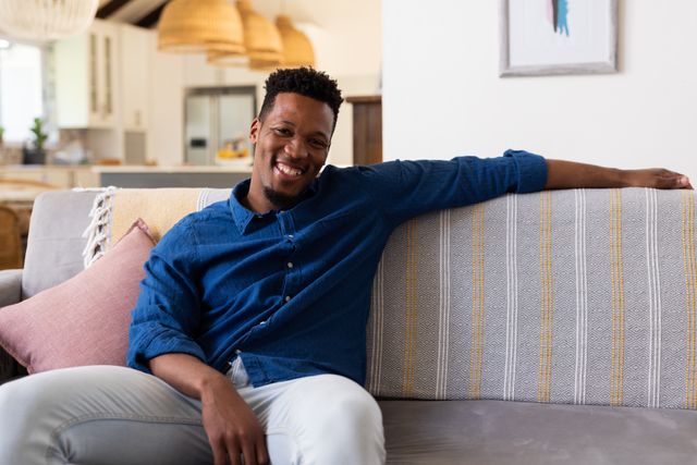 Portrait of happy african american man sitting on couch smiling to camera at home, with copy space. Domestic life and leisure time concept.