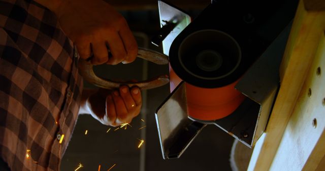 Close-up shows hands sharpening a horseshoe on a grinder, with visible sparks flying. Useful for illustrating concepts such as craftsmanship, metalwork, working with tools, skill, and industrial work. Can be used in articles or projects related to handcrafted items, trade skills, manual labor, and vocational training.