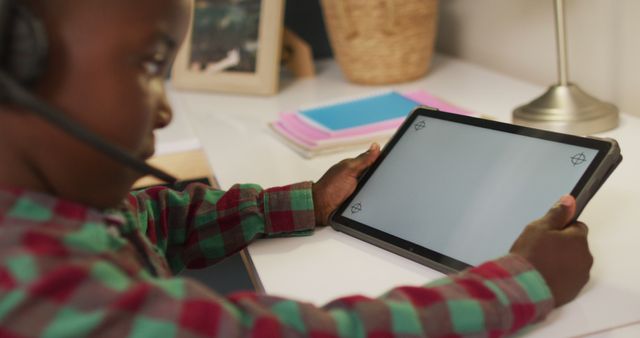 The image shows a young African American boy using a tablet at home. He is wearing a headset and appears to be focusing on a task on the device. This can be used for depicting modern education, digital learning, and study environments involving technology. Ideal for educational materials, tech advertising, online learning platforms, and blog posts about children's education and technology use.