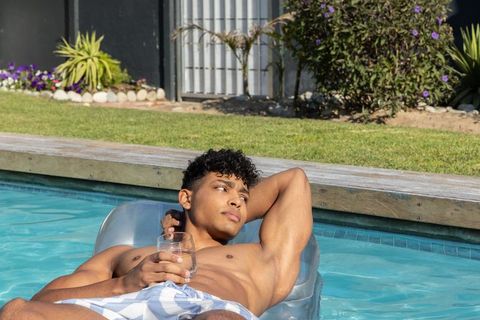 African-american man relaxing on inflatable lounger in sunlit backyard pool holding tumbler