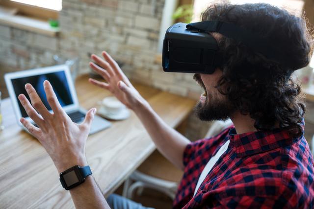 Man enjoying an immersive experience using a VR headset while sitting in a coffee shop. The scene represents modern technology and its integration into everyday life. Perfect for illustrating concepts of digital innovation, VR technology, casual working environments, and tech-savvy lifestyles.