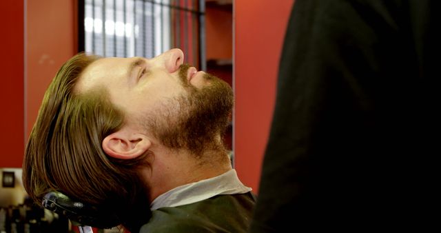 A Caucasian man is getting a haircut or a beard trim at a barbershop, with copy space. His relaxed posture suggests trust in the professional attending to his grooming needs.