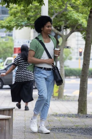 Walking black man carrying coffee on urban sidewalk wearing casual green shirt and jeans