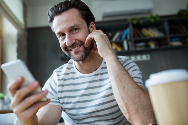 Smiling man holding mobile phone in coffee shop, wearing striped shirt and beard. Ideal for use in advertisements for mobile technology, coffee shops, casual lifestyle, or communication services. Suitable for blog posts, social media content, and marketing materials highlighting modern, relaxed, and connected lifestyles.