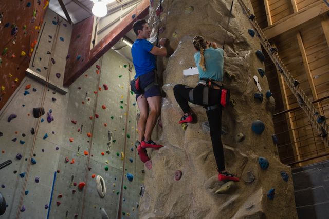 Two athletes are rock climbing on an indoor climbing wall at a fitness studio. They are wearing climbing gear and appear focused on their ascent. This image can be used for promoting fitness centers, climbing gyms, sports training programs, or adventure sports activities. It highlights teamwork, physical fitness, and the thrill of rock climbing.