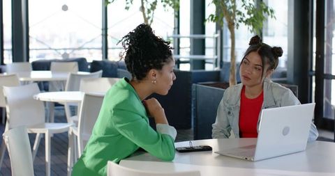 Multicultural women collaborating over laptop and discussing project in modern cafe lounge