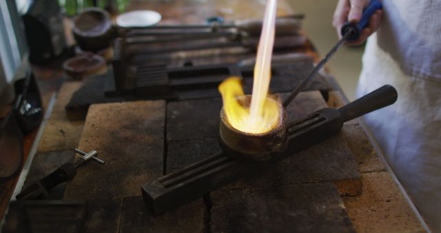 Jeweler meltting metal in crucible with torch in workshop. Suitable for visuals related to skilled craftsman, artisanal jewelry, metalworking techniques, handcrafted items, how-to guides on jewelry making.