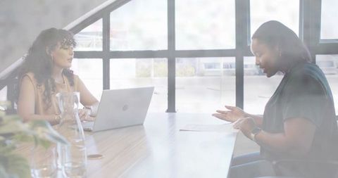 Two women collaborating in sunlit boardroom with laptop, documents and casual office attire