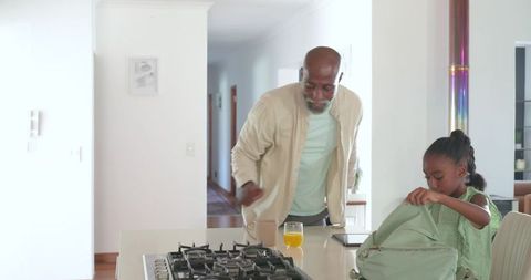 African american grandfather helping granddaughter pack backpack at bright kitchen island