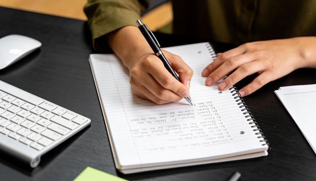 Hands writing in spiral notebook at tidy dark desk holding black pen, keyboard and mouse