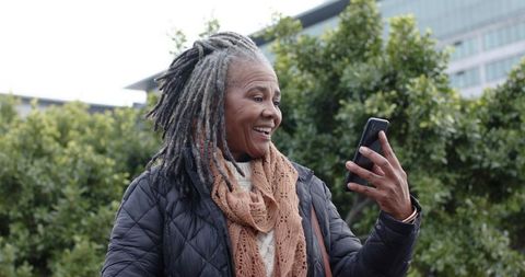 African american senior woman using smartphone outdoors, smiling in urban greenspace